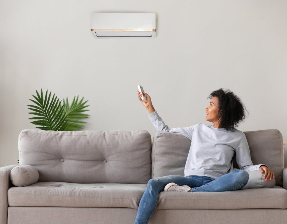 Smiling young African American woman using air conditioner, cooler system remote controller, switching, setting comfort temperature in living room, resting on cozy sofa at home, enjoy fresh air